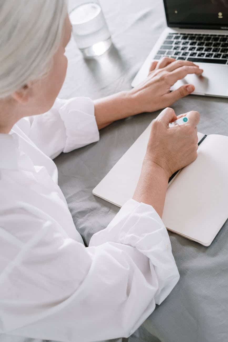 an elderly woman taking notes while using a laptop